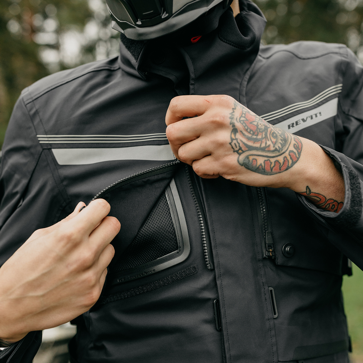 A person with a tattoo on their hand who is zipping up the front pocket of their motorcycle jacket